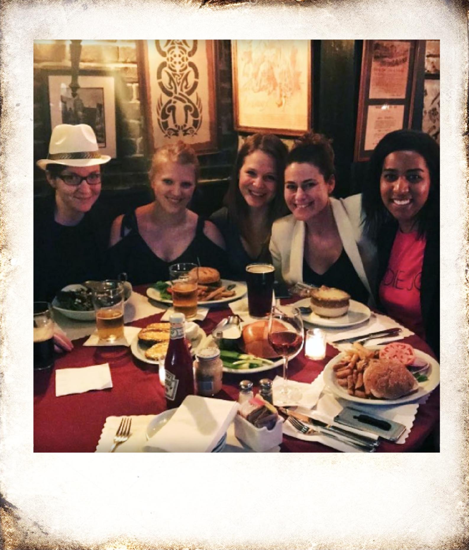 Five women smiling and sitting closely together at a restaurant table with plates of food and drinks including burgers, fries, and beer.